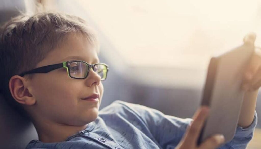 A child wearing glasses, sitting on a couch and looking at a tablet, with a focus on screen use.