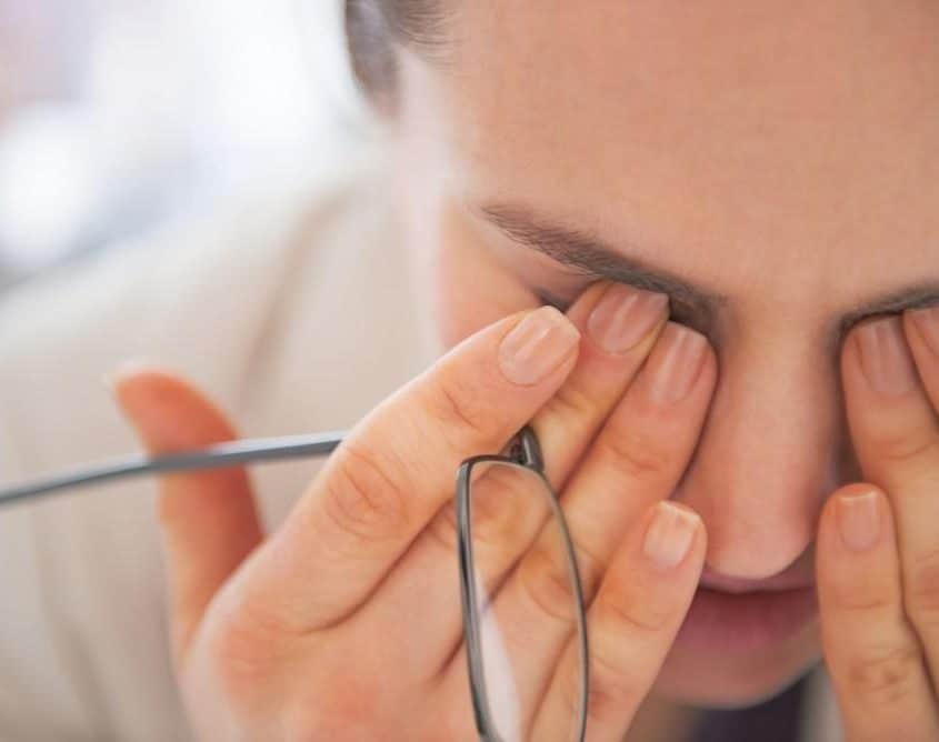 A person rubbing their eyes, possibly experiencing eye strain or dry eyes, while holding a pair of glasses.